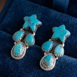 May include: A pair of turquoise and silver earrings. Each earring features a star-shaped turquoise stone at the top, with three additional turquoise stones below. The earrings are set in silver and rest on a blue velvet surface.