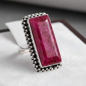 Könnte beinhalten: Silberring mit einem großen, rechteckigen, facettierten Rubin-Edelstein. Der Rubin hat eine tiefe, satte rote Farbe. Der Ring hat einen perlenbesetzten Rand.