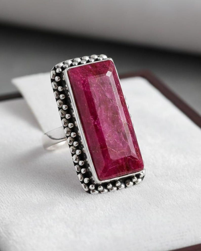 May include: A silver ring featuring a large, rectangular, faceted ruby gemstone. The ruby is a deep, rich red color. The ring has a beaded border around the gemstone.