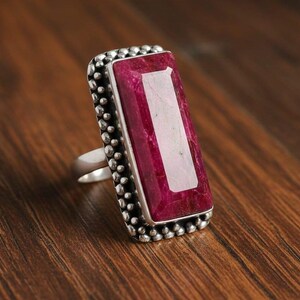 May include: A silver ring with a large, rectangular, ruby-colored gemstone. The gemstone is set in a silver bezel with a dotted border. The ring is displayed on a wooden surface.