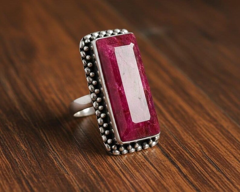 May include: A silver ring with a large, rectangular, ruby-colored gemstone. The gemstone is set in a silver bezel with a dotted border. The ring is displayed on a wooden surface.