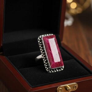 May include: A silver ring with a large, rectangular, ruby-colored gemstone. The gemstone is set in a beaded bezel. The ring is displayed in a wooden jewelry box with a black velvet interior.