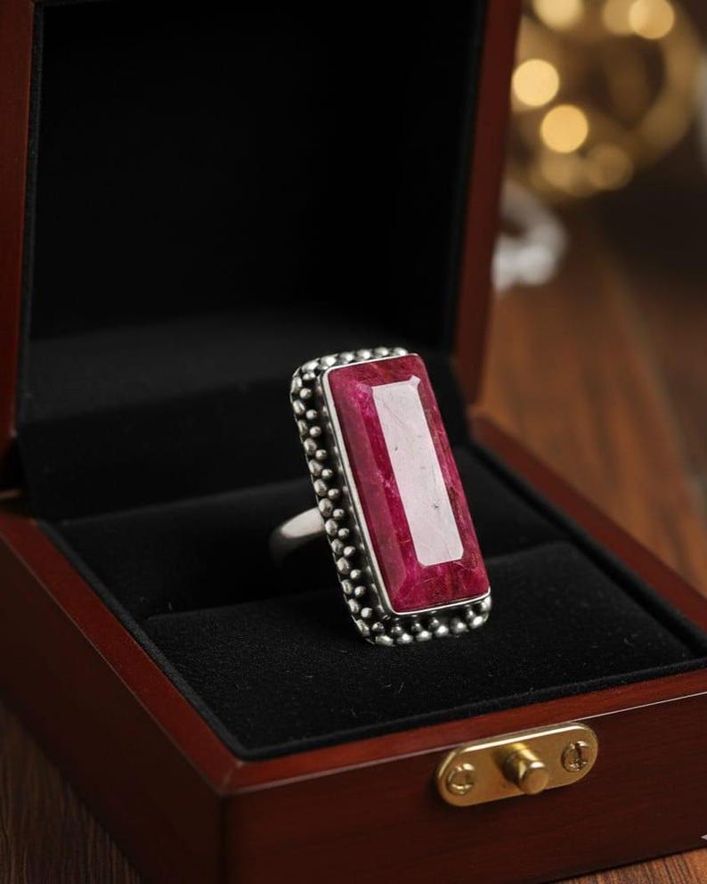May include: A silver ring with a large, rectangular, ruby-colored gemstone. The gemstone is set in a beaded bezel. The ring is displayed in a wooden jewelry box with a black velvet interior.