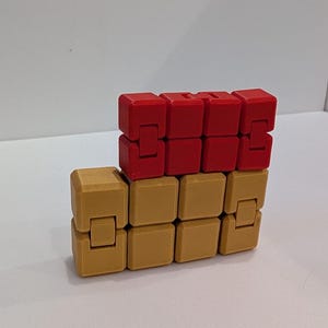 May include: A two-tone fidget cube toy. The top row of cubes is red, and the bottom row is tan. The cubes are connected and can be manipulated to create different shapes. The toy is designed to relieve stress and improve focus.