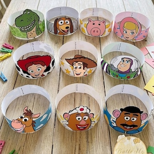 Op de afbeelding: Een set van negen ronde papieren kronen met kleurrijke illustraties van diverse geanimeerde personages, waaronder een groene dinosaurus, een bruine hond, een roze varken, een blonde pop, een cowgirl, een cowboy, een ruimtevaarder en twee aardappelfiguren.