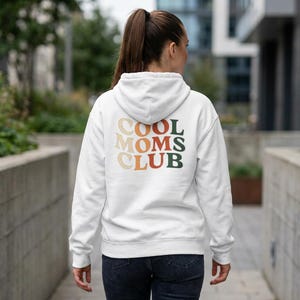 May include: A white hooded sweatshirt with the text "COOL MOMS CLUB" in a retro font on the back. The text is in shades of orange, green, and beige. The sweatshirt is being worn by a person.