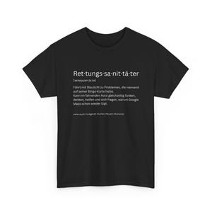 May include: Black t-shirt with white text. The text reads "Ret-tungs-sa-nit-tä-ter" with a German definition below. The definition describes someone who drives with blue lights to problems, and more. The shirt is a crew neck.