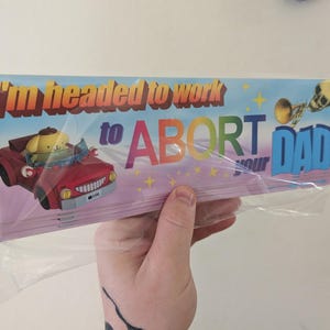 May include: A rectangular bumper sticker in a clear plastic sleeve featuring a yellow cartoon character driving a red car. The text reads I'm headed to work to ABORT your DAD in colorful lettering, accompanied by a small skull and trumpet graphic.
