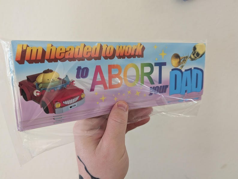 May include: A rectangular bumper sticker in a clear plastic sleeve featuring a yellow cartoon character driving a red car. The text reads I'm headed to work to ABORT your DAD in colorful lettering, accompanied by a small skull and trumpet graphic.