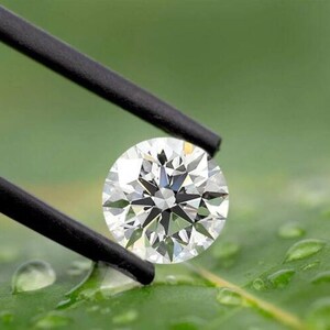 Op de afbeelding: Een briljant geslepen diamant, vastgehouden door een zwarte pincet, tegen een wazige groene achtergrond. De diamant schittert met vele facetten, die het licht reflecteren. De afbeelding is een close-up, die de helderheid van de edelsteen benadrukt.
