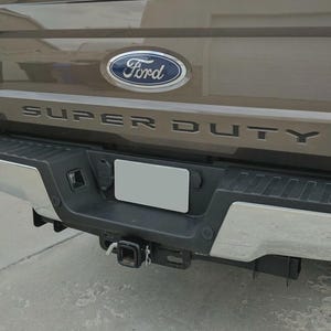 May include: Rear view of a brown Ford Super Duty truck. The Ford logo is centered above the words "SUPER DUTY". The truck has a black bumper with a trailer hitch and a license plate.