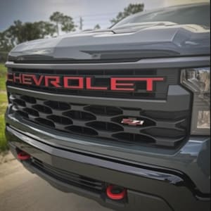 May include: Close-up of a gray truck grille with the red word "CHEVROLET" across the top. The grille has a black honeycomb pattern and a red "Z71" emblem. Red tow hooks are visible on the black bumper.