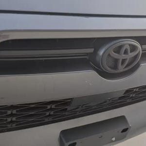 May include: Close-up of a dark gray car grille with a black Toyota emblem. The grille has horizontal and vertical lines, and a black license plate holder is visible at the bottom. The car's front end is in focus.
