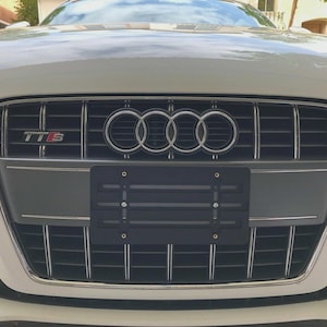 May include: Close-up of a white Audi TT-S car front. The grille features the Audi logo and the letters "TTS". A black license plate holder is mounted on the front bumper.