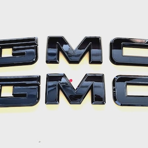 May include: Six glossy black GMC emblems. The letters are in a bold, sans-serif font. The letters are arranged in two rows of three, with the letters G, M, and C visible. The emblems are likely for a vehicle.
