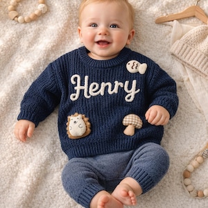 May include: A smiling baby wearing a navy blue sweater with the name "Henry" in white script. The sweater has patches of a fish, mushroom, and hedgehog. The baby is also wearing blue pants and a beige knit hat with a pom-pom.