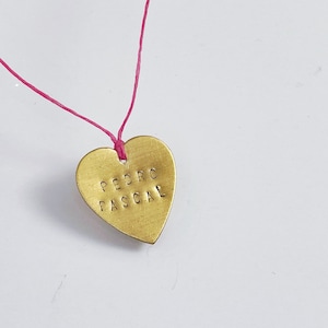 May include: A gold heart-shaped pendant with the name "PEDRO PASCAL" engraved on it. The pendant is attached to a pink string.