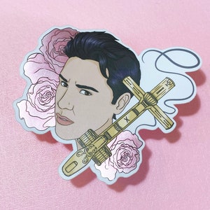 May include: A sticker featuring a man with dark hair and a serious expression. He is holding a gold cross with a floral design. The background is pink with a silver foil effect.