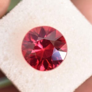 Peut inclure: Une pierre précieuse ronde à facettes d'une couleur rouge profond avec une taille brillant. La pierre présente de multiples facettes triangulaires et en forme de cerf-volant qui reflètent la lumière, créant un motif complexe de tons rouges.
