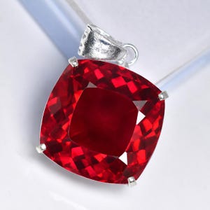 May include: A square-cut, deep red gemstone pendant featuring a faceted surface that reflects light. The stone is secured by four silver-toned prongs and includes a silver-toned metal bail at the top for attachment to a chain or necklace.