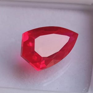 Op de afbeelding: Een gefacetteerde, levendige rode edelsteen met een unieke schildachtige vorm. De edelsteen heeft een lichtgekleurd, bijna wit, midden en is geplaatst tegen een witte achtergrond. De slijping van de steen creëert meerdere reflecterende oppervlakken.