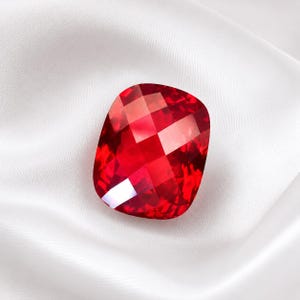 Puede incluir: Una gema de color rojo intenso y vibrante con forma de cojín y una superficie facetada que refleja la luz. La piedra presenta un aspecto brillante y transparente con bordes geométricos definidos y un acabado pulido, mostrando un tono carmesí rico y saturado.