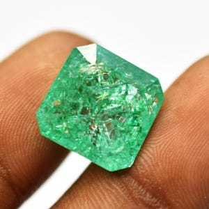 Könnte beinhalten: Ein facettierter, smaragdgrüner Edelstein mit geometrischer Form. Der Edelstein hat eine transluzente Qualität mit sichtbaren inneren Einschlüssen und einer hellen, reflektierenden Oberfläche. Der Stein wird zwischen Fingern gehalten.