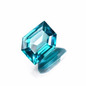 May include: A faceted, teal-colored gemstone with a rectangular shape and beveled edges. The gem is set against a white background, with a shadow cast below. The gem's facets reflect light, creating a sparkling effect.