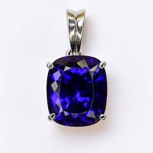 Può includere: Ciondolo con pietra preziosa blu scuro a taglio quadrato con anello argentato. La pietra preziosa è sfaccettata, riflettendo la luce. Il ciondolo è incastonato in una montatura in metallo argentato con quattro griffe che tengono la pietra. Misura circa 2 cm.