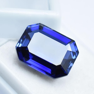 Può includere: Una gemma sfaccettata, taglio rettangolare, di colore blu intenso. La gemma ha una superficie luminosa e riflettente e un centro chiaro e rettangolare. I bordi sono ben definiti, mostrando la precisione del taglio. La gemma è su sfondo bianco.