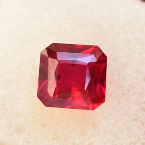 May include: A rectangular-cut ruby gemstone with multiple facets. The gem is a deep red colour, reflecting light. The gemstone is set against a neutral, off-white background.