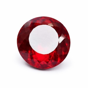 Puede incluir: Una piedra preciosa redonda y facetada de color rojo intenso y vibrante. La piedra presenta un corte circular con numerosas facetas triangulares que reflejan la luz, creando una apariencia brillante y centelleante en su superficie.