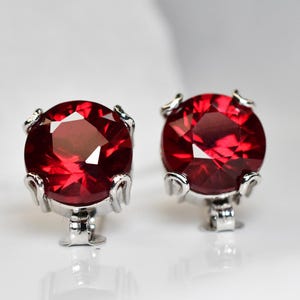 Puede incluir: Un par de pendientes de botón redondos con piedras de color rojo intenso facetadas, engastadas en garras de metal plateado pulido. Cada pendiente presenta un corte circular con múltiples superficies reflectantes.