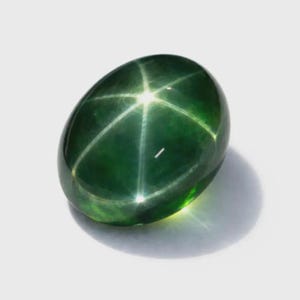 Pode incluir: Uma pedra preciosa oval polida, de cor verde escura. A gema apresenta um padrão estelar de seis raios em sua superfície, criado pela reflexão da luz nas inclusões internas. A pedra está sobre um fundo neutro.
