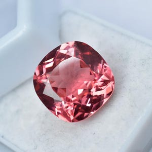 Puede incluir: Una gema facetada, de talla cojín, con un tono rojizo-rosado. La gema se encuentra sobre un fondo blanco, mostrando su intrincada estructura interna y superficies reflectantes. La forma de la gema es cuadrada con esquinas redondeadas.