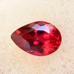 May include: A faceted, pear-shaped, red gemstone. The gem has a high degree of clarity and brilliance, with multiple facets reflecting light. The stone is set against a neutral, off-white background.