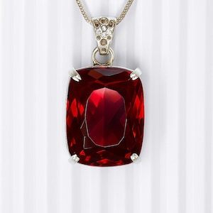 Op de afbeelding: Een zilveren hangerketting met een grote, gefacetteerde, rechthoekige, dieprode edelsteen. De edelsteen wordt op zijn plaats gehouden door vier zilveren pootjes. De ketting is zilverkleurig en de hanger is versierd met kleine, heldere stenen.