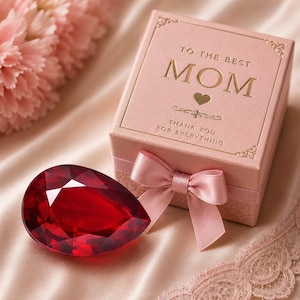 Pode incluir: Uma caixa de presente quadrada rosa claro com um laço de cetim, apresentando texto dourado que diz TO THE BEST MOM e THANK YOU FOR EVERYTHING, acompanhada por uma grande pedra preciosa vermelha escura facetada em forma de pera sobre tecido.