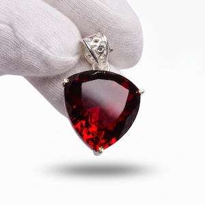 May include: A triangular pendant featuring a deep red faceted gemstone set in a silver-toned metal frame. The setting includes a decorative, openwork bail at the top, designed to hold a chain, with three small prongs securing the stone in place.