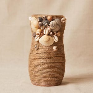 以下が含まれることがあります： 天然の麻ひもで包まれた装飾的な花瓶で、さまざまな貝殻のクラスターで飾られています。 花瓶は円筒形で、貝殻は上部に集中しています。 カラーパレットはニュートラルで、茶色とベージュの色合いです。