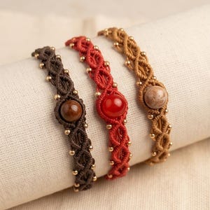 May include: Three macrame bracelets featuring intricate knotted cord patterns in dark brown, bright red, and tan. Each bracelet is centred with a single smooth round gemstone bead and accented with small metallic gold beads woven into the cord design.