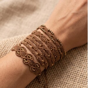 May include: A set of five brown macrame bracelets featuring intricate woven patterns and small metallic gold-toned beads integrated into the cord. The bracelets are layered together, showcasing varying knot designs and textures in a uniform brown colour.
