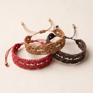 May include: Three adjustable bracelets featuring a braided cord design in red, tan, and dark brown. Each bracelet has a woven pattern and thin pull strings finished with small gold-toned metal beads at the ends for a secure and customisable fit.