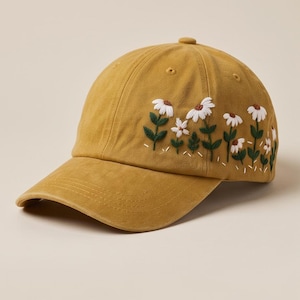 May include: A mustard yellow baseball cap featuring embroidered white daisy flowers with green stems and leaves along the side panel. The cap has a curved brim, a button on top, and stitched eyelets for ventilation, showcasing a detailed floral design.