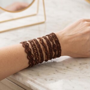May include: A wide brown macramé bracelet featuring intricate knotted patterns and small metallic beads woven throughout the design. The bracelet wraps multiple times around the wrist, creating a layered appearance with a consistent dark brown cord.