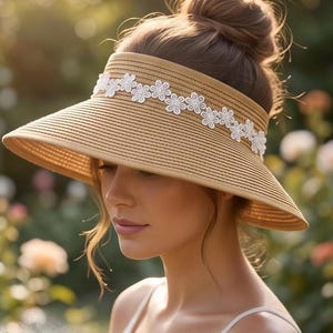 May include: A tan straw visor featuring a wide, curved brim and a decorative band of white embroidered flowers across the front. The visor has a textured, woven appearance and is designed to provide shade with its structured, open-top construction.