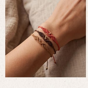 May include: Three macramé bracelets in coral, dark brown, and tan, each featuring a braided cord design with a small, centred gold-toned metallic bead. The bracelets are thin, adjustable, and have delicate cord ends with tiny gold-toned beads.