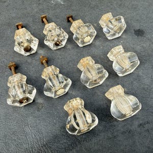 May include: A collection of nine clear glass cabinet knobs featuring a faceted, hexagonal shape. Each knob is attached to a threaded metal bolt with a visible patina, showing signs of age and oxidation on the hardware components.