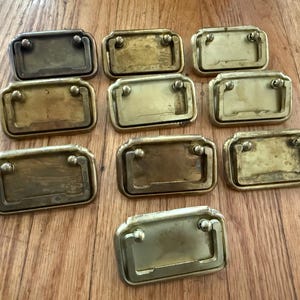 May include: A collection of ten rectangular brass drawer pulls, each featuring a hinged drop handle attached to a flat backplate. The metal hardware displays varying degrees of aged patina, ranging from bright polished gold to dark bronze tones.