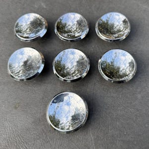May include: Seven circular buttons with a highly polished, reflective silver-tone finish. Each button features a smooth, domed surface that mirrors the surrounding environment, including visible reflections of trees and a bright, cloudy blue sky.